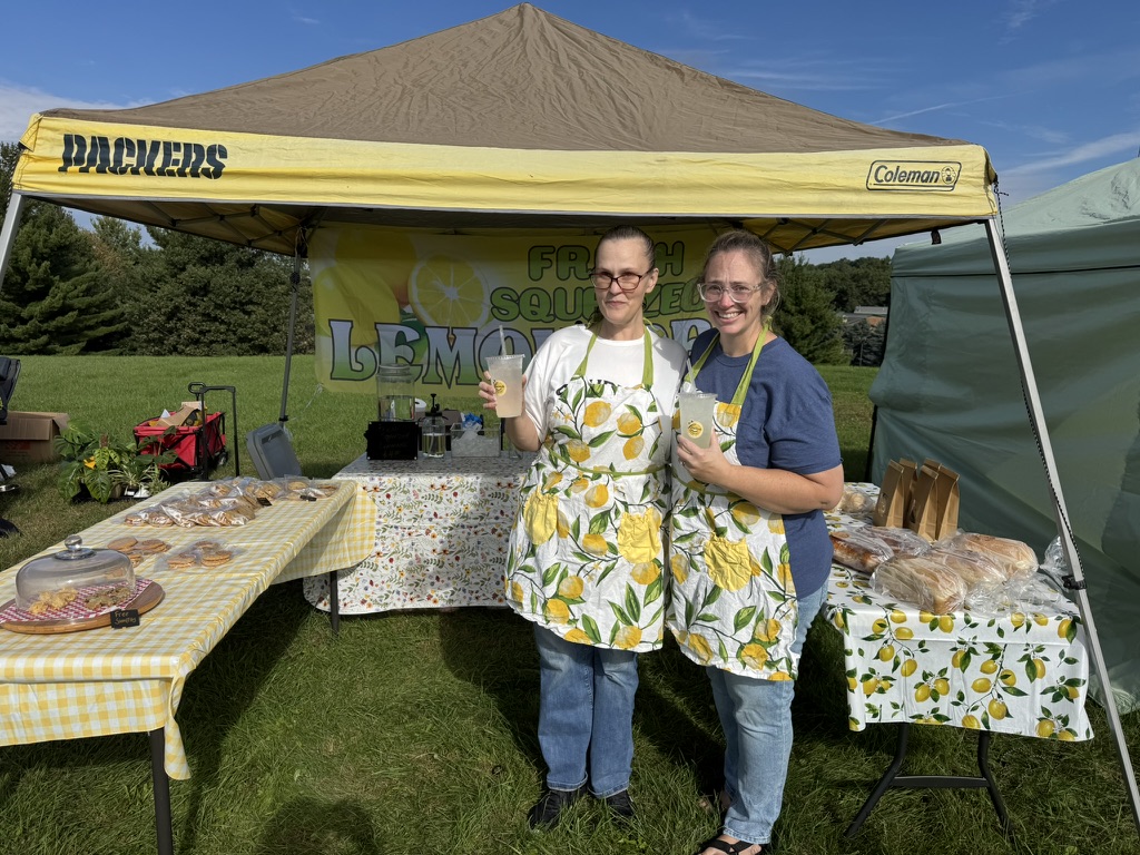 Rachel and Maggie holding lemonade together at their outdoor stand